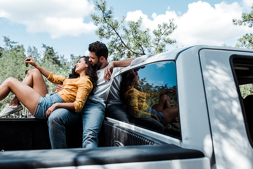 low angle view of handsome bearded man sitting on car and looking at happy young woman