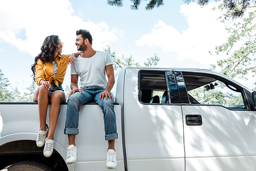 low angle view of handsome bearded man sitting on car and looking at happy woman