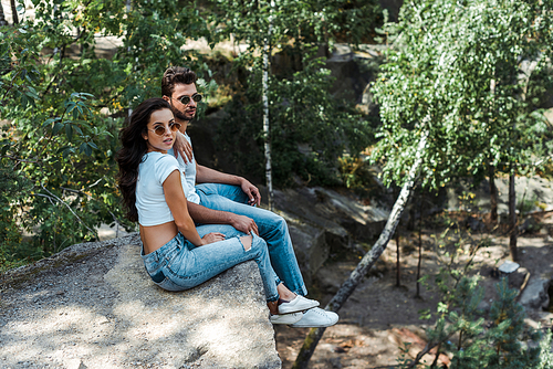 handsome man and attractive woman in sunglasses sitting near trees