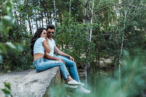 selective focus of handsome man and attractive woman in sunglasses sitting near trees