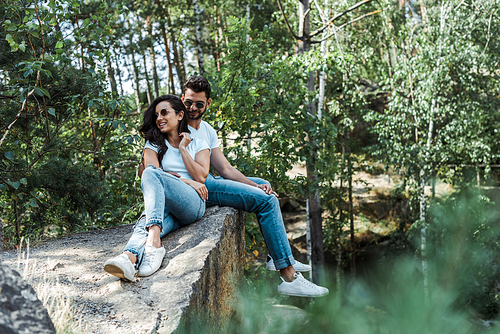 selective focus of handsome man and cheerful woman in sunglasses sitting near trees with leaves