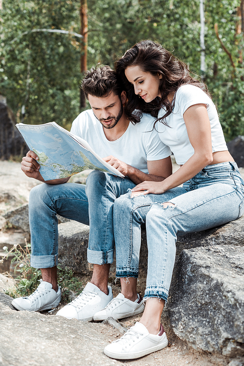 handsome man looking at map near attractive girl sitting on stone