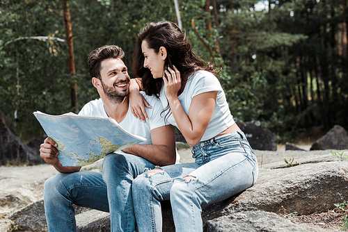 happy man holding map near cheerful girl sitting on stone