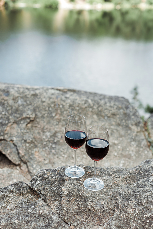 selective focus of wine glasses with red aromatic wine on stone near lake