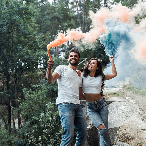 attractive woman and cheerful bearded man holding smoke bombs outside