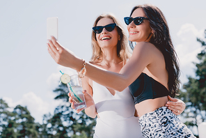 low angle view of happy brunette and blonde women in swimsuits taking selfie