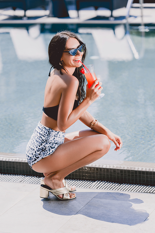 happy sexy brunette woman in swimsuit and sunglasses sitting near swimming pool and drinking cocktail