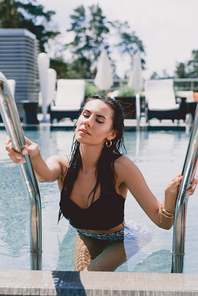 wet brunette woman with closed eyes and hands on metal handrails getting out from water in swimming pool
