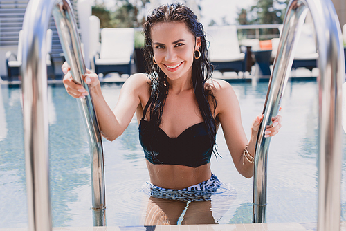 selective focus of happy wet brunette woman with hands on metal handrails getting out from water in swimming pool