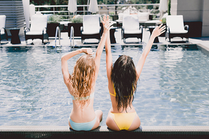 back view of blonde and brunette wet girls sitting near swimming pool with hands in air