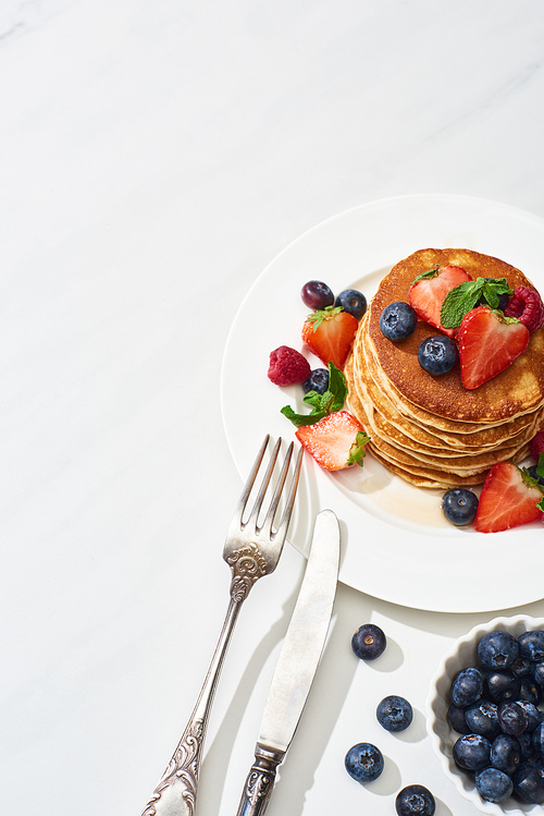 top view of delicious pancakes with maple syrup, blueberries and strawberries on plate near fork and knife on marble white surface