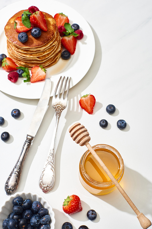 top view of delicious pancakes with honey, blueberries and strawberries on plate near fork and knife on marble white surface