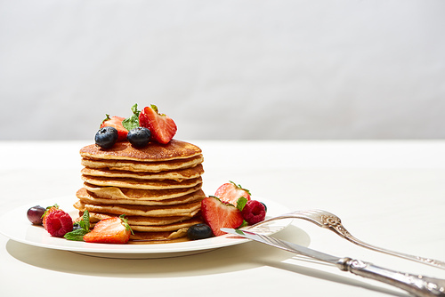 delicious pancakes with blueberries and strawberries on plate with cutlery on white surface isolated on grey