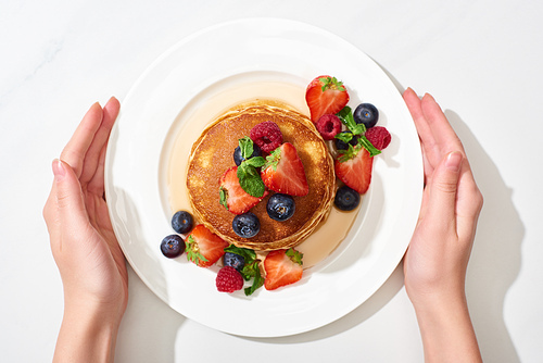 partial view of woman holding plate with delicious pancakes with honey, blueberries and strawberries on marble white surface