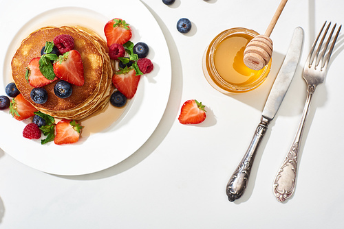 top view of delicious pancakes with honey, blueberries and strawberries on plate near cutlery on marble white surface