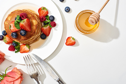 top view of delicious pancakes with honey, blueberries and strawberries on plate near cutlery and rose on marble white surface