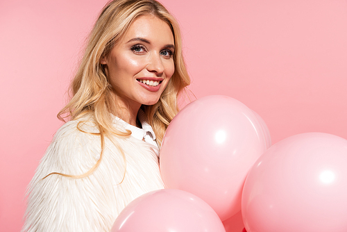 smiling beautiful blonde woman in faux fur jacket holding pink balloons isolated on pink