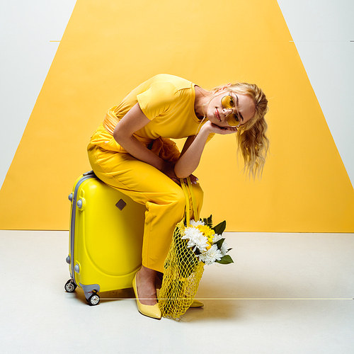 pretty blonde girl sitting on luggage and holding string bag with flowers on white and yellow