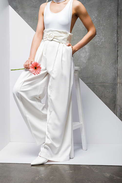 cropped view of woman posing with hand in pocket near chair with pink flower on white and grey