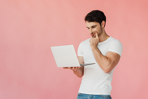 pensive muscular man in white t-shirt using laptop isolated on pink