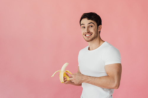 smiling muscular man in white t-shirt holding banana and  isolated on pink