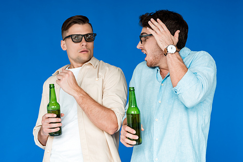 two worried friends in 3d glasses holding bottles of beer and looking at each other isolated on blue