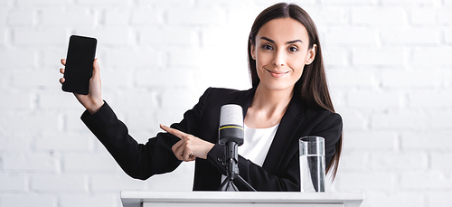 beautiful smiling lecturer pointing with finger at smartphone with blank screen while standing on podium tribune and 