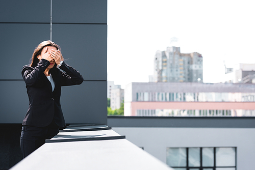 young businesswoman, suffering from fear of heights, standing on rooftop and covering face with hands