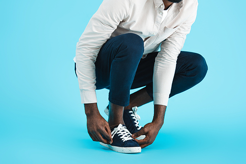 cropped view of african american man tying up laces on sneakers isolated on blue