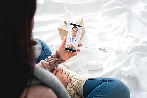 cropped view of ill woman with pills and thermometer having online consultation with male doctor on smartphone