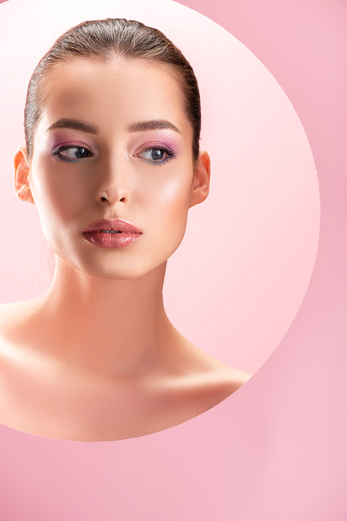 portrait of beautiful naked woman with shiny makeup looking through round paper hole isolated on pink