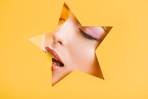 portrait of beautiful woman with shiny makeup in paper star shaped hole with closed eyes and open mouth isolated on yellow