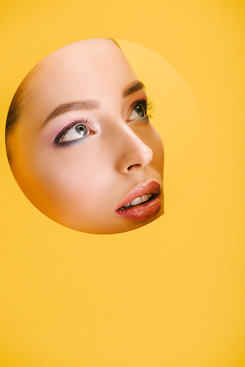 portrait of beautiful woman with shiny makeup in paper round hole looking away isolated on yellow