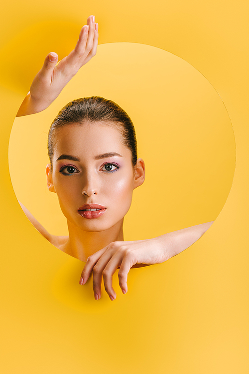 portrait of beautiful woman with shiny makeup in paper round hole with hands isolated on yellow