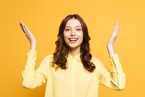 excited girl showing wow gesture with raised hands while smiling at camera isolated on yellow