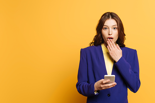 shocked girl covering mouth with hand while holding smartphone on yellow background