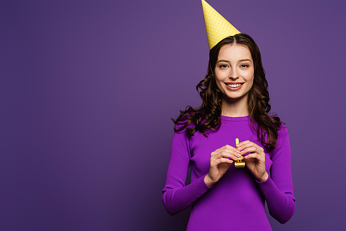 happy girl in party cap holding party horn while smiling at camera on purple background