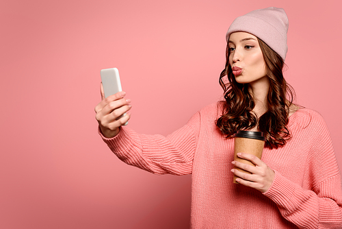 attractive girl with duck face expression taking selfie while holding cofee to go on pink background