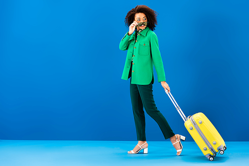 smiling african american woman holding sunglasses and holding travel bag on blue background