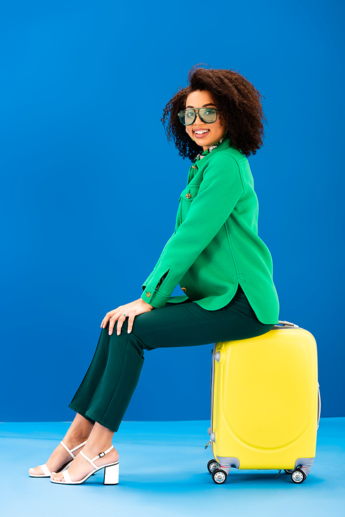 smiling african american woman sitting on travel bag on blue background