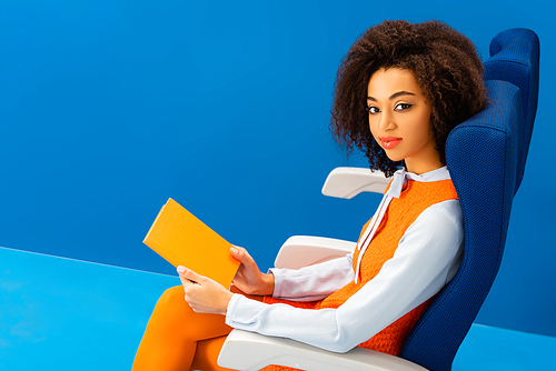 high angle view of smiling african american in retro dress holding book on blue background