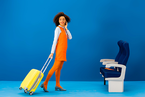 smiling african american in retro dress walking with travel bag on blue background