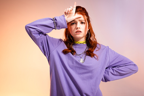 redhead female teenager in bad mood showing loser sign, on purple and beige