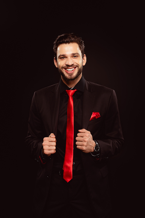 Handsome man in black suit and red tie smiling at camera isolated on black