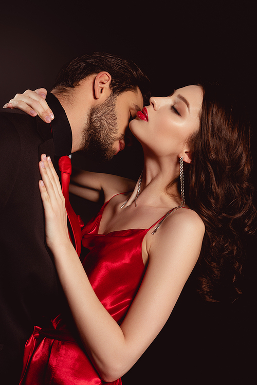Side view of man in suit kissing attractive woman in red dress isolated on black