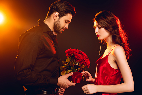 Side view of elegant man giving roses and gift box to beautiful woman in red dress on black background with lighting