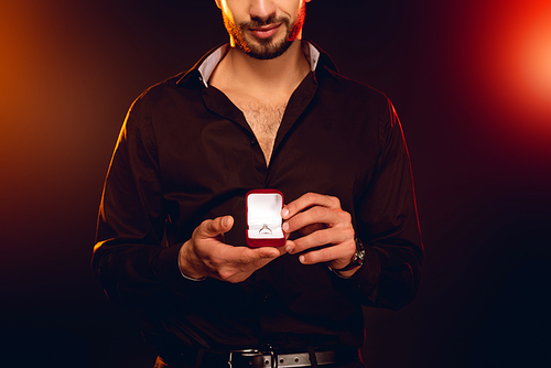 Cropped view of elegant man holding gift box with jewelry ring on black background with lighting