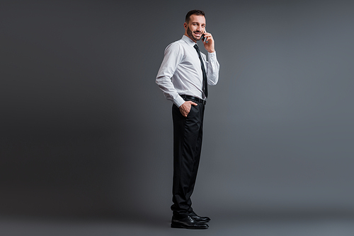 smiling businessman in suit talking on smartphone while standing with hand in pocket on grey