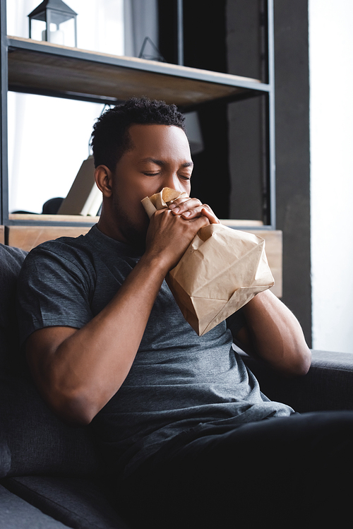 Worried african american man breathing with paper bag while having panic attack at home