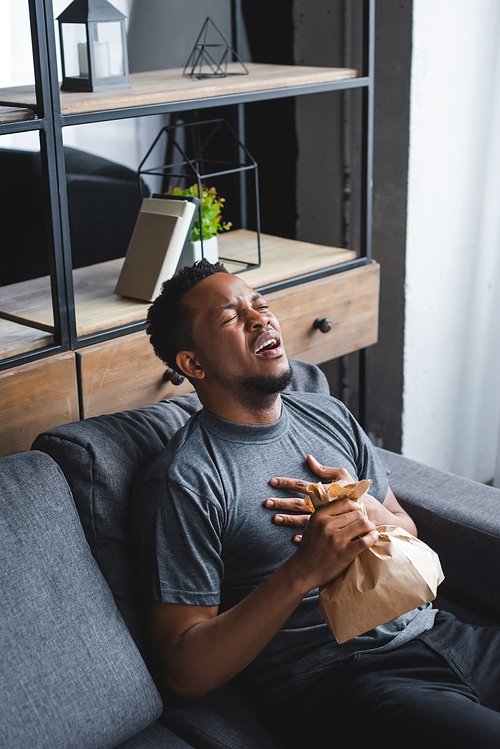 stressed african american man holding paper bag while having panic attack at home